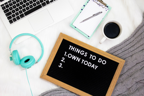 working at home with laptop headphones coffee and a sign that lists things to do today own today.